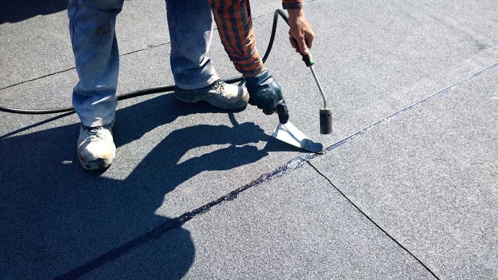 A homeowner examines a ceiling stain beneath a roof leak while a bucket catches dripping water.