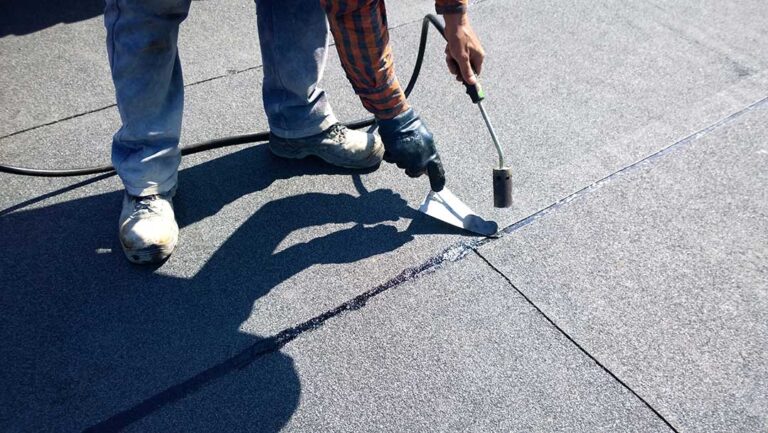 A homeowner examines a ceiling stain beneath a roof leak while a bucket catches dripping water.
